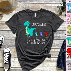 Op de afbeelding: Donkergrijs t-shirt met een dinosaurusontwerp. Het shirt heeft de tekst "DADDYSAURUS" en "LIKE A NORMAL DAD, BUT MORE AWESOME." Een ontwerptabel met 12 dinosaurusontwerpen staat rechts.