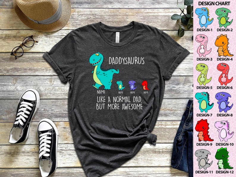 Op de afbeelding: Donkergrijs t-shirt met een dinosaurusontwerp. Het shirt heeft de tekst "DADDYSAURUS" en "LIKE A NORMAL DAD, BUT MORE AWESOME." Een ontwerptabel met 12 dinosaurusontwerpen staat rechts.