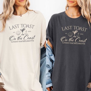 May include: Two white t-shirts with a black graphic design. The graphic design says "Last Toast est. 2024 On the Coast Linda's Bachelorette" with a seashell, cocktail glass, and anchor. The t-shirts are shown on models wearing denim jackets. The left t-shirt is ivory and the right t-shirt is pepper.