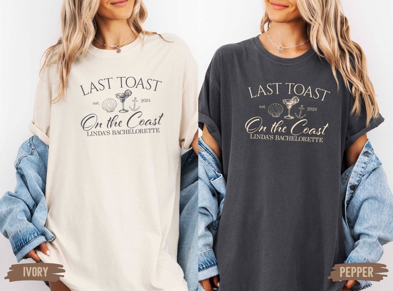 May include: Two white t-shirts with a black graphic design. The graphic design says "Last Toast est. 2024 On the Coast Linda's Bachelorette" with a seashell, cocktail glass, and anchor. The t-shirts are shown on models wearing denim jackets. The left t-shirt is ivory and the right t-shirt is pepper.