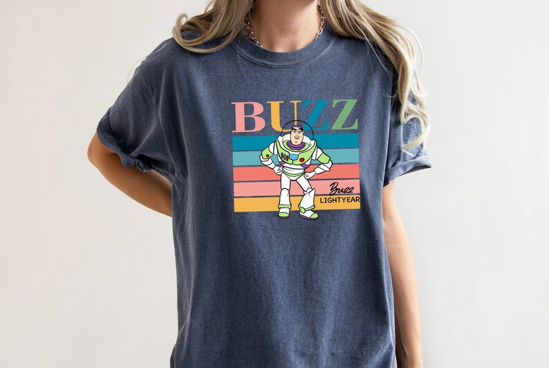 May include: A navy blue t-shirt with a retro-style graphic of Buzz Lightyear from Toy Story. The graphic features Buzz standing with his arms on his hips, and the text "Buzz Lightyear" is printed below him.
