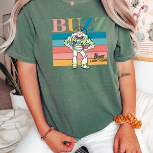May include: Green t-shirt with a retro-style graphic of Buzz Lightyear from Toy Story. The graphic features Buzz in his space ranger suit with the text "Buzz Lightyear" underneath.