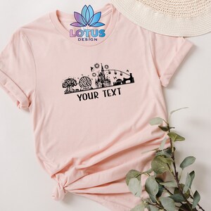K&ouml;nnte beinhalten: Ein pinkfarbenes T-Shirt mit einer schwarzen Silhouette eines Schlosses und anderer Disney-Parkattraktionen. Der Text "LOTUS DESIGN" und "YOUR TEXT" ist auf dem T-Shirt aufgedruckt.