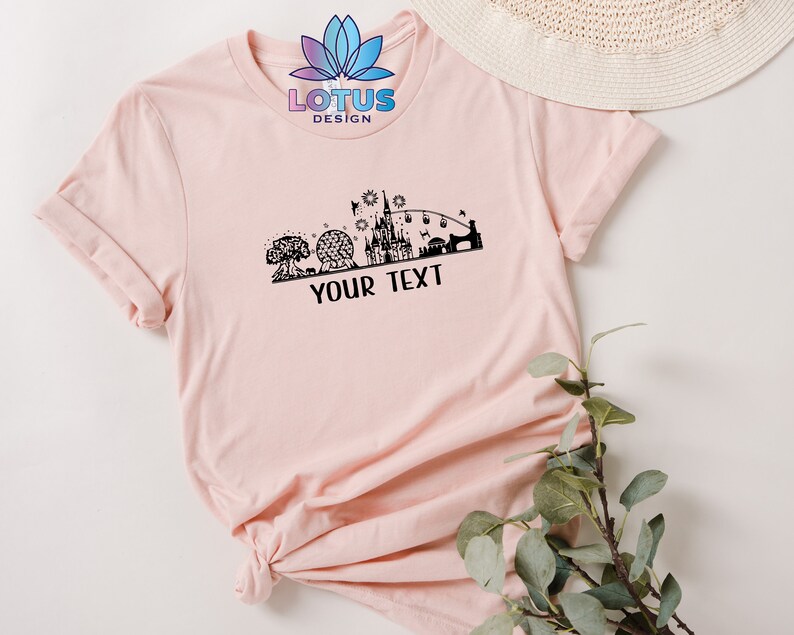 K&ouml;nnte beinhalten: Ein pinkfarbenes T-Shirt mit einer schwarzen Silhouette eines Schlosses und anderer Disney-Parkattraktionen. Der Text "LOTUS DESIGN" und "YOUR TEXT" ist auf dem T-Shirt aufgedruckt.