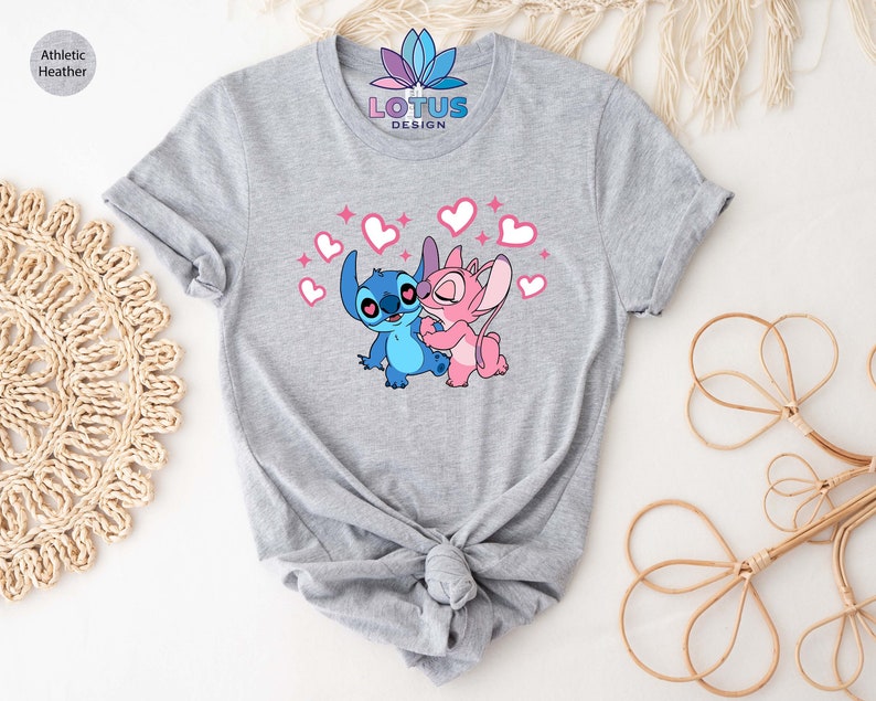 K&ouml;nnte beinhalten: Graues T-Shirt mit einem Motiv von Stitch und Angel aus Lilo & Stitch. Die Figuren umarmen sich und sind von rosa Herzen umgeben. Der Text "LOTUS DESIGN" ist auf dem T-Shirt aufgedruckt.