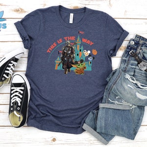May include: A navy blue t-shirt featuring a graphic of a Mandalorian character and a Baby Yoda character in front of a castle. The text "This is the way" is printed on the shirt.