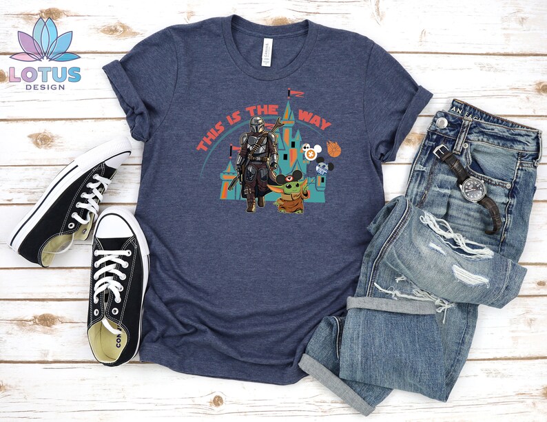 May include: A navy blue t-shirt featuring a graphic of a Mandalorian character and a Baby Yoda character in front of a castle. The text "This is the way" is printed on the shirt.