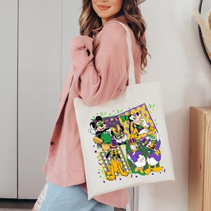 Puede incluir: Un bolso tote de color crema con un gr&aacute;fico colorido de Mickey Mouse, Minnie Mouse, Goofy, Donald Duck y Pluto con atuendos de Mardi Gras. El dise&ntilde;o incluye confeti y formas geom&eacute;tricas moradas, verdes y amarillas. El bolso tiene un dise&ntilde;o simple y resistente.