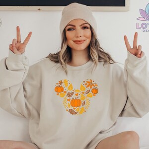 May include: A beige sweatshirt with a Mickey Mouse-shaped design featuring fall leaves and pumpkins in orange and yellow colors. The text "SAND" is visible in the top left corner of the image.