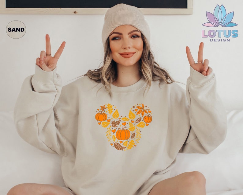 May include: A beige sweatshirt with a Mickey Mouse-shaped design featuring fall leaves and pumpkins in orange and yellow colors. The text "SAND" is visible in the top left corner of the image.