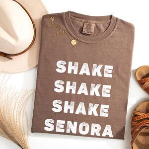 May include: Brown t-shirt with the text "Shake Shake Shake Senora" in white lettering. The shirt is folded and laid flat on a white surface.