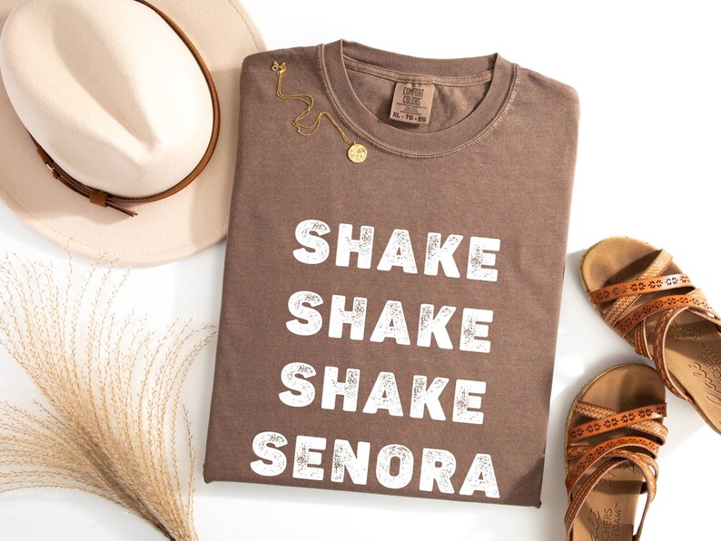May include: Brown t-shirt with the text "Shake Shake Shake Senora" in white lettering. The shirt is folded and laid flat on a white surface.