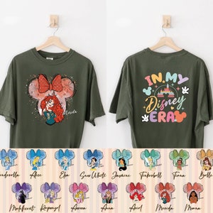 Puede incluir: Una camiseta verde con un gr&aacute;fico de una princesa de Disney con un lazo en la cabeza. La parte posterior de la camiseta dice "In My Disney Era" con un gr&aacute;fico del castillo de Disney. Hay 12 im&aacute;genes m&aacute;s peque&ntilde;as de diferentes princesas de Disney con lazos en la cabeza.