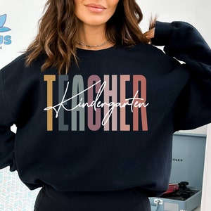K&ouml;nnte beinhalten: Ein schwarzer Pullover mit dem Text "TEACHER Kindergarten" in bunten vertikalen Streifen. Der Text ist in einer modernen, handschriftlichen Schrift.