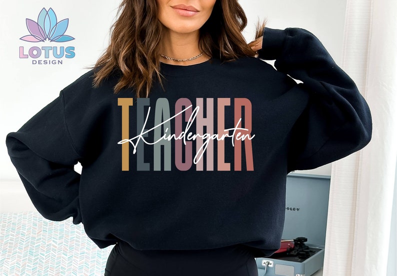K&ouml;nnte beinhalten: Ein schwarzer Pullover mit dem Text "TEACHER Kindergarten" in bunten vertikalen Streifen. Der Text ist in einer modernen, handschriftlichen Schrift.