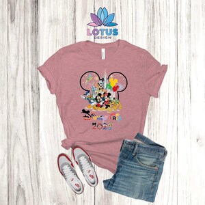 Puede incluir: Una camiseta rosa con un contorno negro de oreja de Mickey Mouse. Dentro del contorno est&aacute;n Mickey Mouse y otros personajes de Disney, incluyendo Donald Duck, Goofy y Pluto. El texto "Disney Trip 2023" est&aacute; impreso debajo de los personajes.
