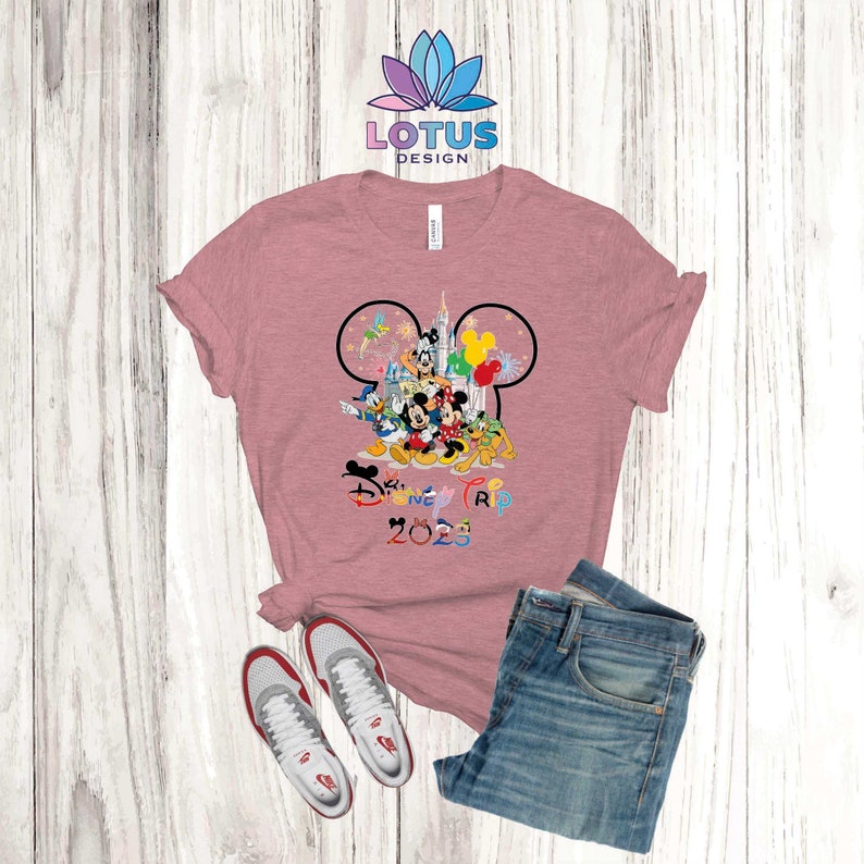 Puede incluir: Una camiseta rosa con un contorno negro de oreja de Mickey Mouse. Dentro del contorno est&aacute;n Mickey Mouse y otros personajes de Disney, incluyendo Donald Duck, Goofy y Pluto. El texto "Disney Trip 2023" est&aacute; impreso debajo de los personajes.
