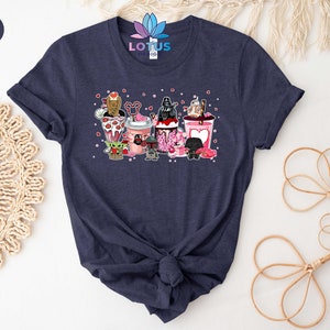 May include: A heather navy blue t-shirt with a graphic of Star Wars characters in pink Valentine's Day themed cups. The characters include Yoda, Darth Vader, Chewbacca, and R2-D2.
