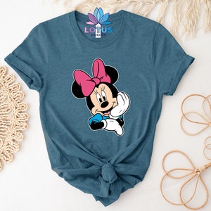K&ouml;nnte beinhalten: Ein blaues T-Shirt mit einem Cartoon-Bild von Minnie Mouse. Minnie Mouse tr&auml;gt eine rote Schleife und ein blaues Kleid. Sie l&auml;chelt und winkt.