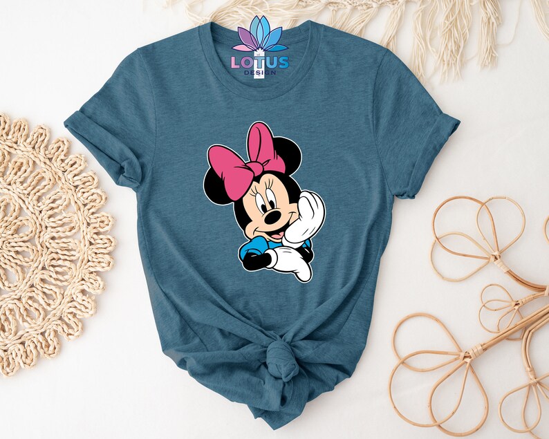 K&ouml;nnte beinhalten: Ein blaues T-Shirt mit einem Cartoon-Bild von Minnie Mouse. Minnie Mouse tr&auml;gt eine rote Schleife und ein blaues Kleid. Sie l&auml;chelt und winkt.