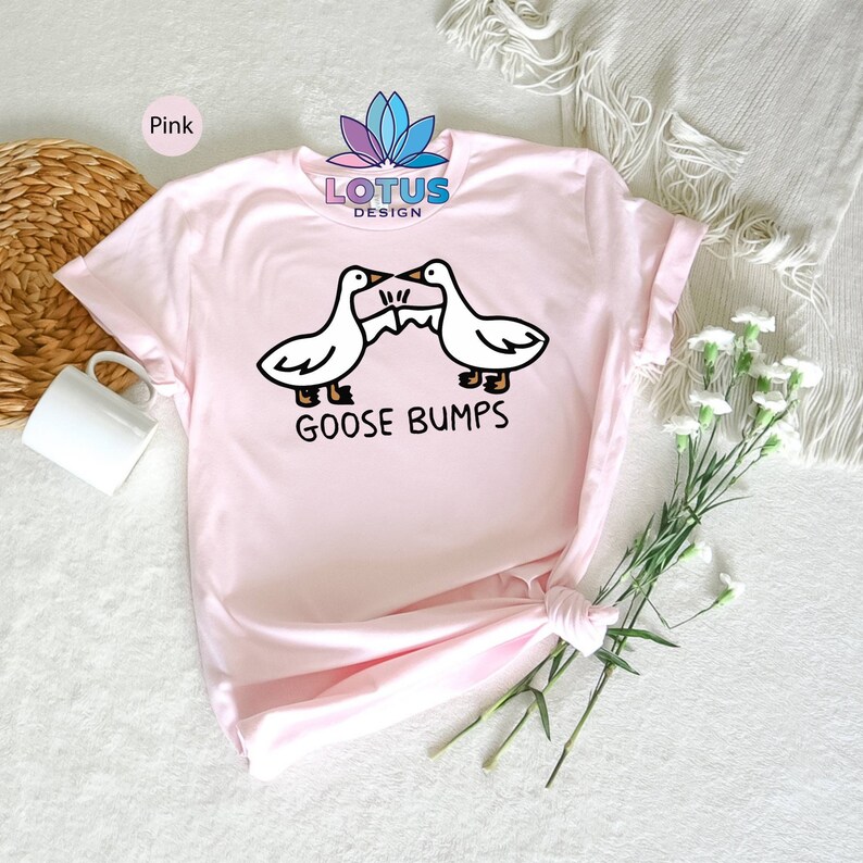Op de afbeelding: Roze T-shirt met een afbeelding van twee cartoon ganzen die elkaar aankijken met de tekst "GOOSE BUMPS" eronder.