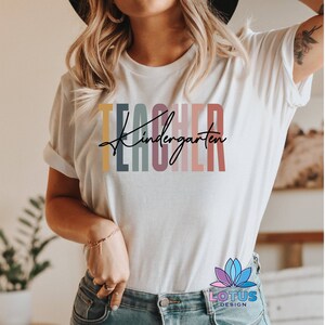 K&ouml;nnte beinhalten: Ein wei&szlig;es T-Shirt mit dem Text "Kindergarten Teacher" in bunten, vertikalen Streifen. Der Text ist in einer modernen, handschriftlichen Schrift.