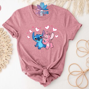 K&ouml;nnte beinhalten: Ein T-Shirt in Heather Mauve mit einem Motiv von Stitch und Angel aus Lilo & Stitch. Das Motiv zeigt Stitch und Angel, die sich mit Herzen und Glitzer umarmen. Der Text "LOTUS DESIGN" ist auf das T-Shirt gedruckt.