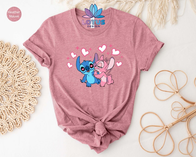 K&ouml;nnte beinhalten: Ein T-Shirt in Heather Mauve mit einem Motiv von Stitch und Angel aus Lilo & Stitch. Das Motiv zeigt Stitch und Angel, die sich mit Herzen und Glitzer umarmen. Der Text "LOTUS DESIGN" ist auf das T-Shirt gedruckt.