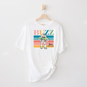 May include: White t-shirt with a colorful retro design featuring Buzz Lightyear from Toy Story. The design includes the words "Buzz" and "Buzz Lightyear" in a colorful font.