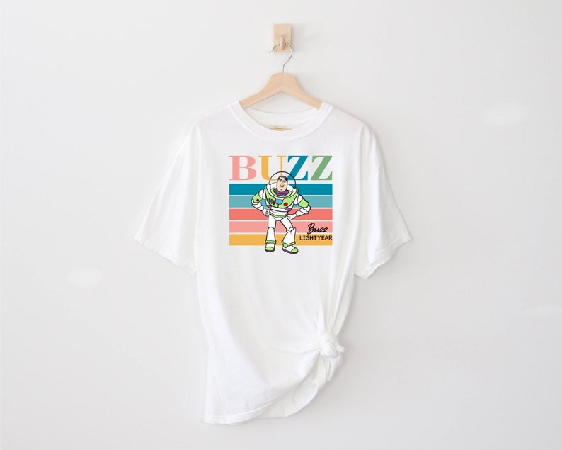 May include: White t-shirt with a colorful retro design featuring Buzz Lightyear from Toy Story. The design includes the words "Buzz" and "Buzz Lightyear" in a colorful font.