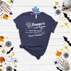 May include: A navy blue t-shirt with a white graphic of a child care center called "Krueger's Little Dreamers". The graphic includes a cartoon of Freddy Krueger, the fictional character from the Nightmare on Elm Street film series. The text on the graphic reads "Child Care Center", "100 Elm Street-Springfield, Illinois", and "Sweet Dreams!"