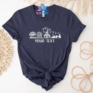 K&ouml;nnte beinhalten: Ein marineblaues T-Shirt mit einer wei&szlig;en Silhouette einer Themenpark-Skyline, einschlie&szlig;lich eines Schlosses, einer Kugel und eines Baumes. Der Text "YOUR TEXT" ist unter der Skyline gedruckt.
