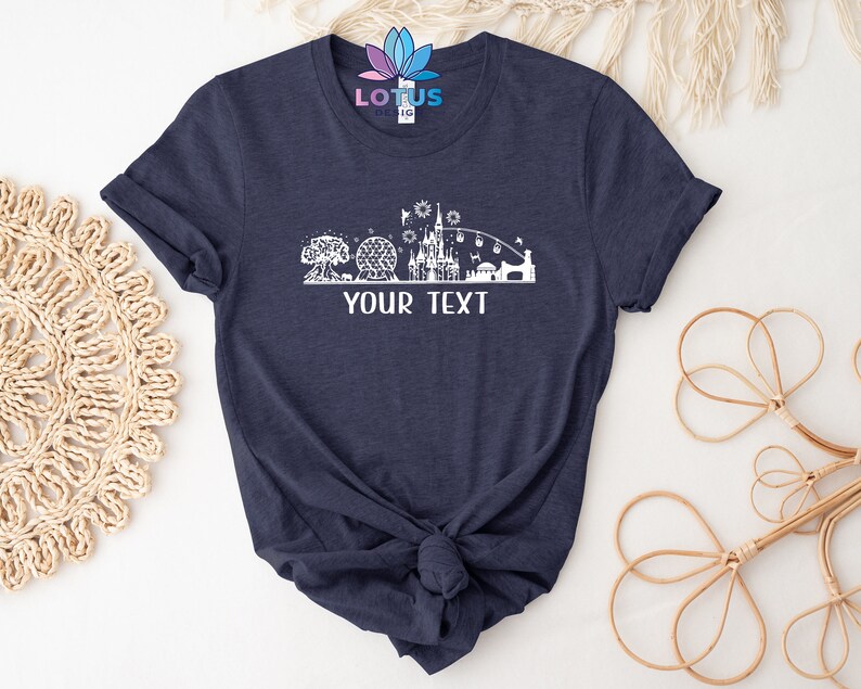 K&ouml;nnte beinhalten: Ein marineblaues T-Shirt mit einer wei&szlig;en Silhouette einer Themenpark-Skyline, einschlie&szlig;lich eines Schlosses, einer Kugel und eines Baumes. Der Text "YOUR TEXT" ist unter der Skyline gedruckt.