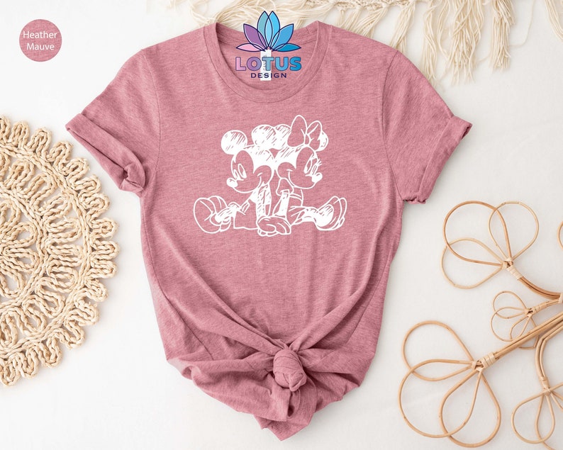 Pode incluir: Uma camiseta de cor malva heather com um contorno branco do Mickey Mouse e da Minnie Mouse de m&atilde;os dadas. O texto "LOTUS DESIGN" est&aacute; impresso na camiseta.