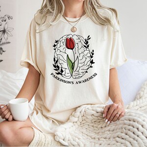 May include: A white t-shirt featuring a black and white illustration of a brain with a red tulip growing from it. The text "Parkinson's Awareness" is printed beneath the illustration.