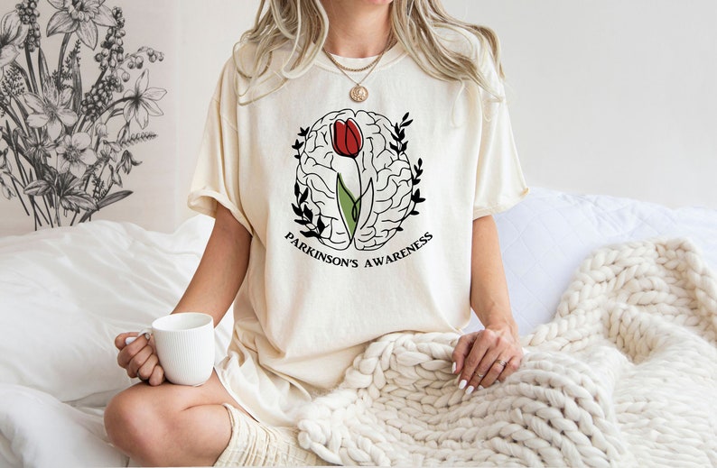 May include: A white t-shirt featuring a black and white illustration of a brain with a red tulip growing from it. The text "Parkinson's Awareness" is printed beneath the illustration.