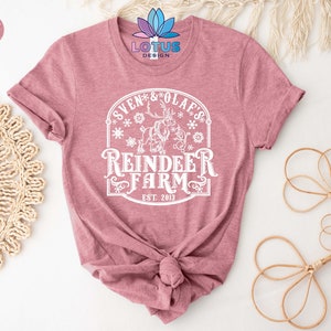 May include: A heather mauve t-shirt with a white graphic design featuring a reindeer and the text "Sven & Olaf's Reindeer Farm Est. 2013".