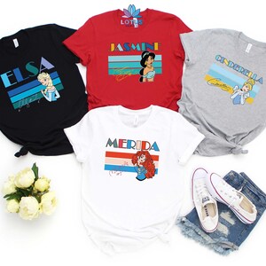 May include: Four t-shirts featuring different Disney princesses with retro-style graphics. The shirts are black, red, grey, and white. Each shirt features a princess's name and a colourful striped design. The princesses featured are Elsa, Jasmine, Cinderella, and Merida.