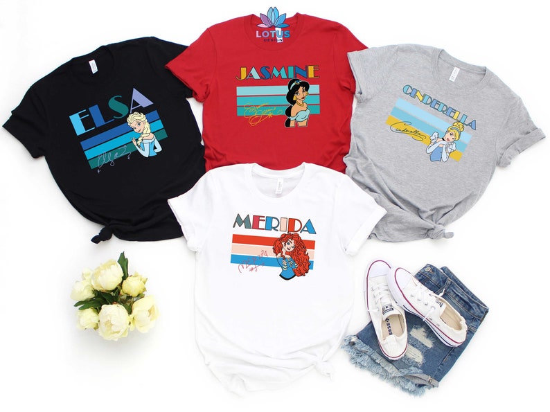 May include: Four t-shirts featuring different Disney princesses with retro-style graphics. The shirts are black, red, grey, and white. Each shirt features a princess's name and a colourful striped design. The princesses featured are Elsa, Jasmine, Cinderella, and Merida.