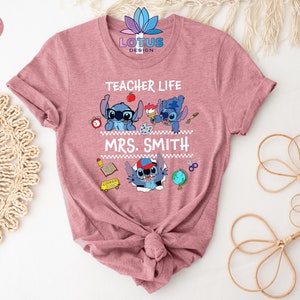 May include: A pink t-shirt with the text "Teacher Life" and a cartoon Stitch character. The text "Mrs. Smith" is below the Stitch character. The shirt is decorated with school supplies like a book, pencil, scissors, and a globe.