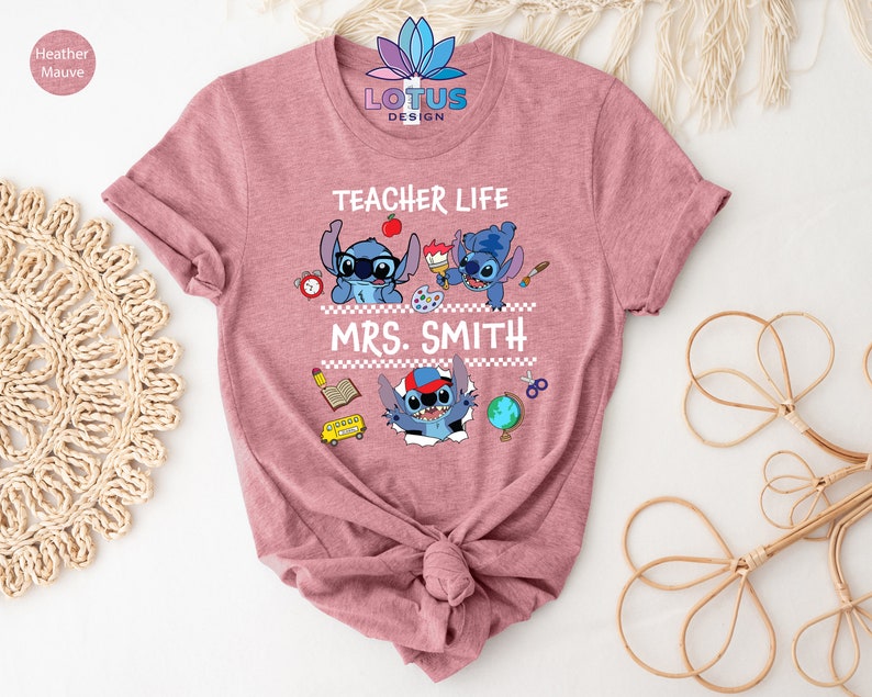 May include: A pink t-shirt with the text "Teacher Life" and a cartoon Stitch character. The text "Mrs. Smith" is below the Stitch character. The shirt is decorated with school supplies like a book, pencil, scissors, and a globe.