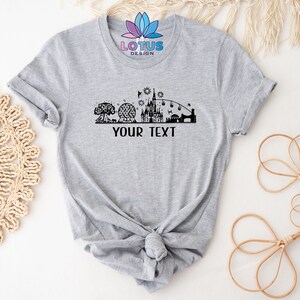 K&ouml;nnte beinhalten: Graues T-Shirt mit einer schwarzen Silhouette einer Disney-Park-Skyline, einschlie&szlig;lich des Schlosses, Spaceship Earth und des Baumes des Lebens. Der Text "YOUR TEXT" befindet sich unterhalb der Skyline.