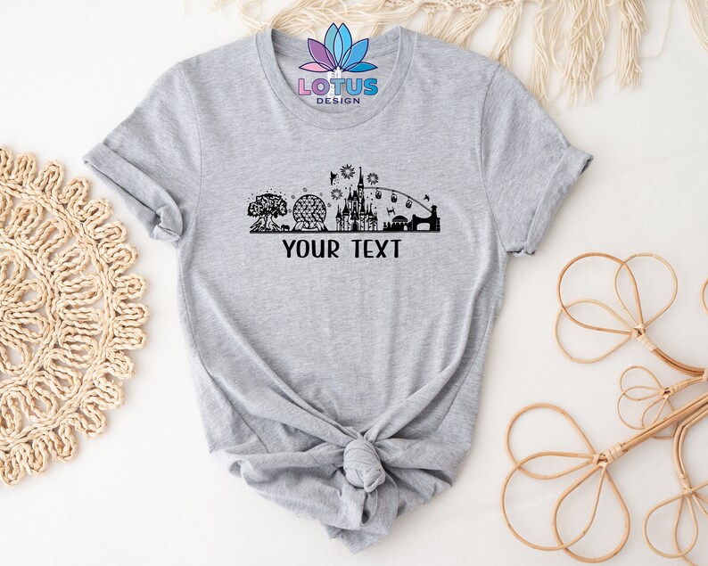 K&ouml;nnte beinhalten: Graues T-Shirt mit einer schwarzen Silhouette einer Disney-Park-Skyline, einschlie&szlig;lich des Schlosses, Spaceship Earth und des Baumes des Lebens. Der Text "YOUR TEXT" befindet sich unterhalb der Skyline.