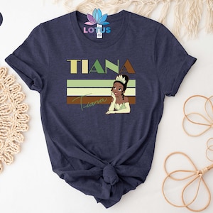 May include: A navy blue t-shirt featuring a retro-style graphic of Tiana, a Disney princess, with the text "Tiana" in bold letters above her.