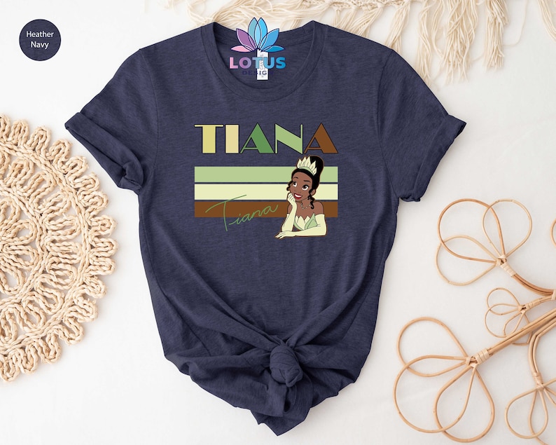 May include: A navy blue t-shirt featuring a retro-style graphic of Tiana, a Disney princess, with the text "Tiana" in bold letters above her.