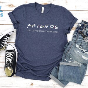 Puede incluir: Una camiseta azul marino con las palabras "FRIENDS" en una fuente estilizada y el texto "Don't let friends fight cancer alone" debajo. La camiseta est&aacute; colocada sobre una superficie de madera blanca con un par de zapatillas negras y un par de pantalones vaqueros azules.