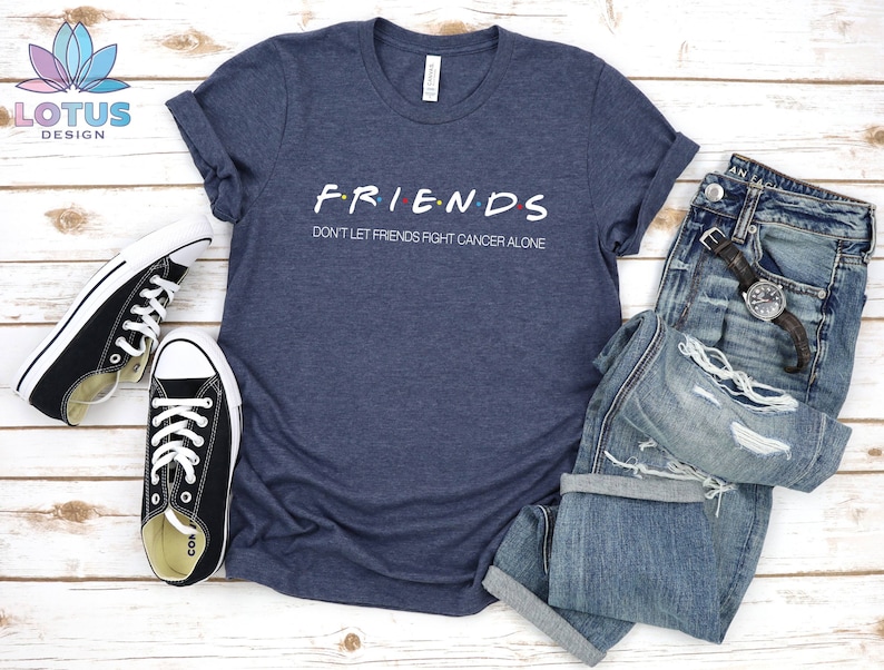 Puede incluir: Una camiseta azul marino con las palabras "FRIENDS" en una fuente estilizada y el texto "Don't let friends fight cancer alone" debajo. La camiseta est&aacute; colocada sobre una superficie de madera blanca con un par de zapatillas negras y un par de pantalones vaqueros azules.