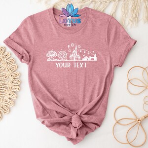 K&ouml;nnte beinhalten: Ein pinkfarbenes T-Shirt mit einer wei&szlig;en Silhouette eines Schlosses und anderer Attraktionen. Der Text "YOUR TEXT" ist unterhalb der Silhouette gedruckt.