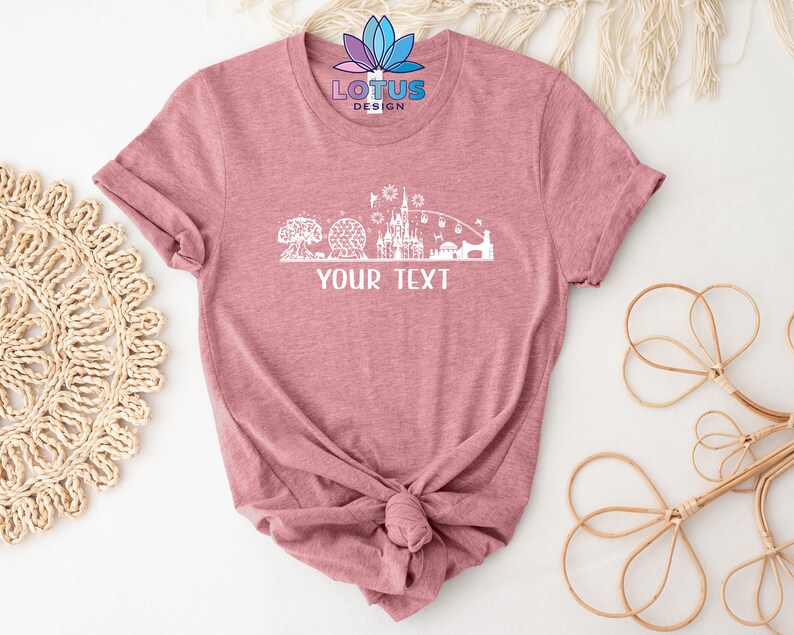 K&ouml;nnte beinhalten: Ein pinkfarbenes T-Shirt mit einer wei&szlig;en Silhouette eines Schlosses und anderer Attraktionen. Der Text "YOUR TEXT" ist unterhalb der Silhouette gedruckt.