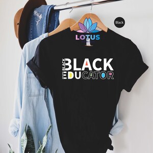 May include: Black t-shirt with the text "I'm Black Educator" in white letters with a rainbow color scheme. The shirt is hanging on a wooden hanger.